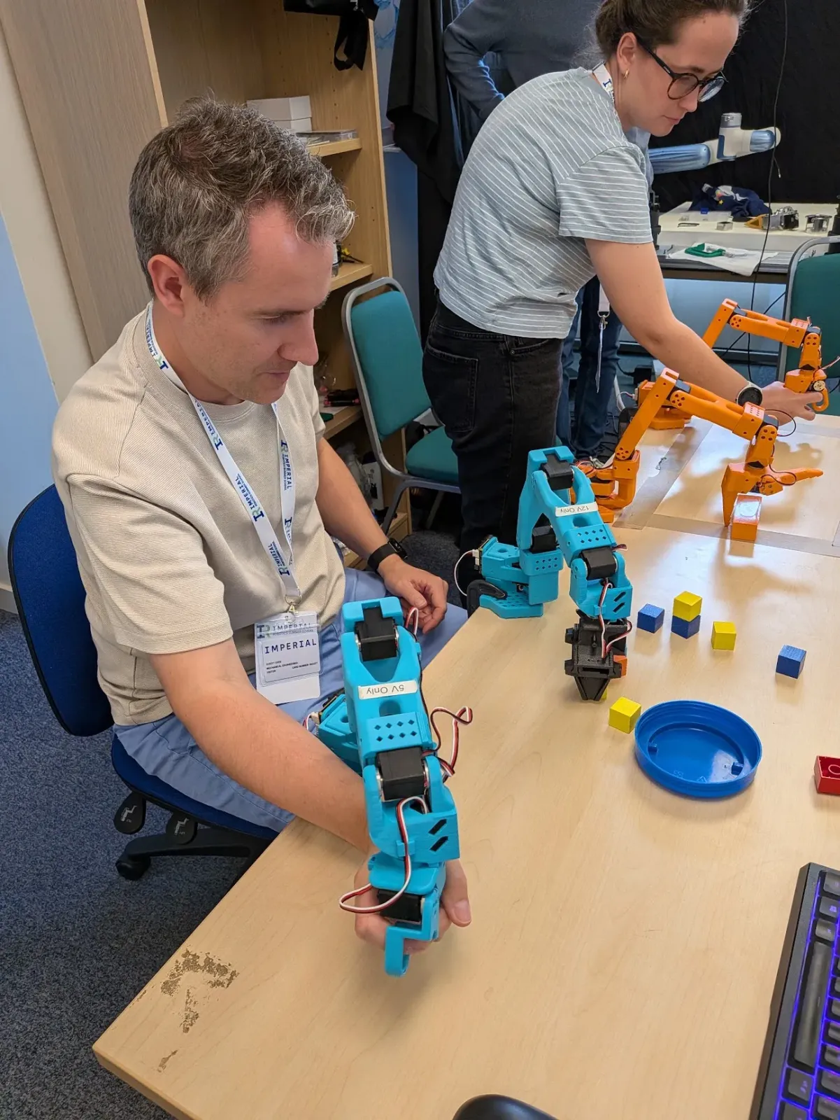Anthony at a lab bench moving a hand-held blue 3D-printed leader arm while a matching arm on the table follows, with coloured blocks to sort and orange arms behind.