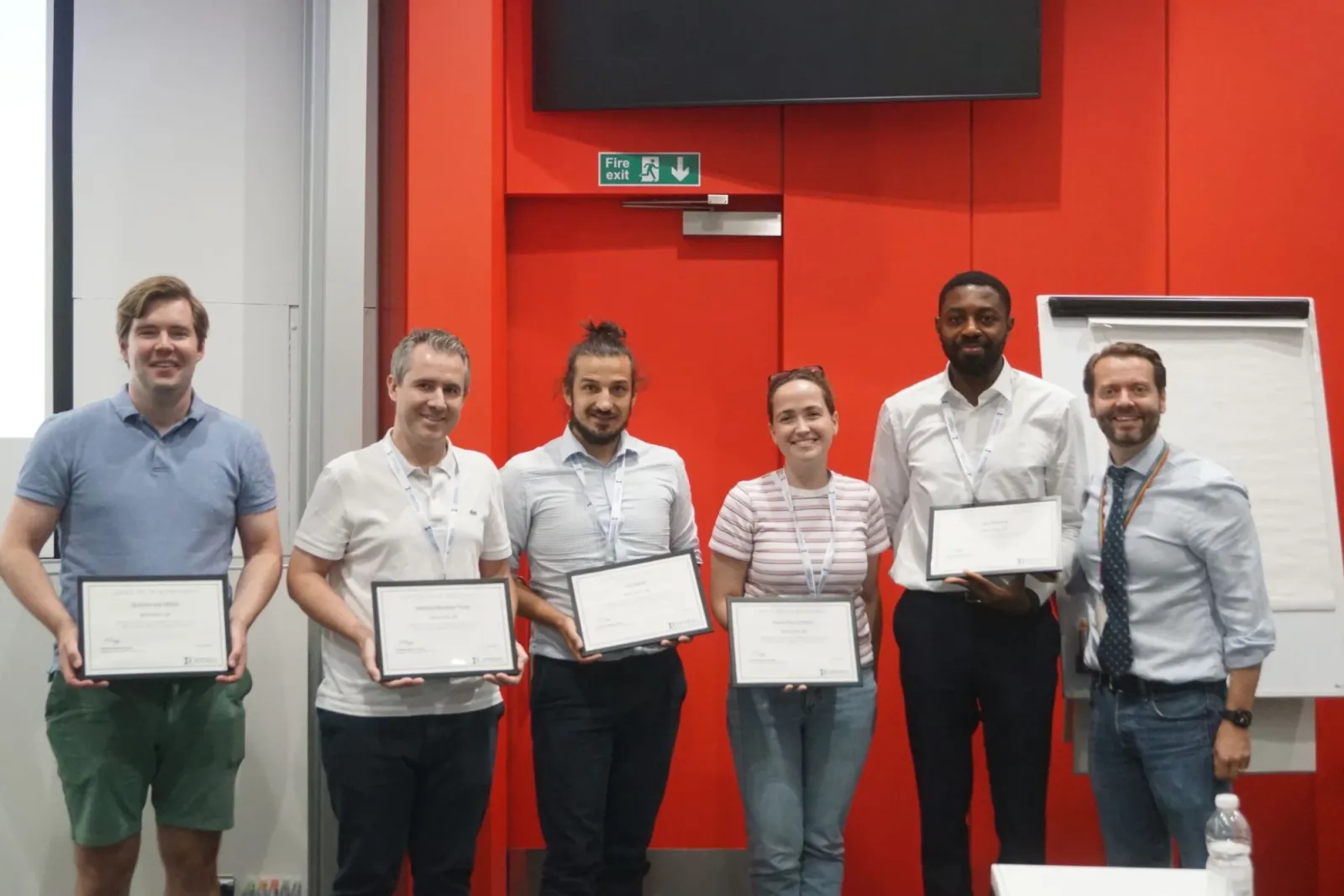 Anthony and four teammates holding framed certificates in front of a red wall, alongside a member of the summer school team.